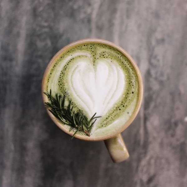 Overhead view of a green matcha latte with heart-shaped foam art and a rosemary sprig.