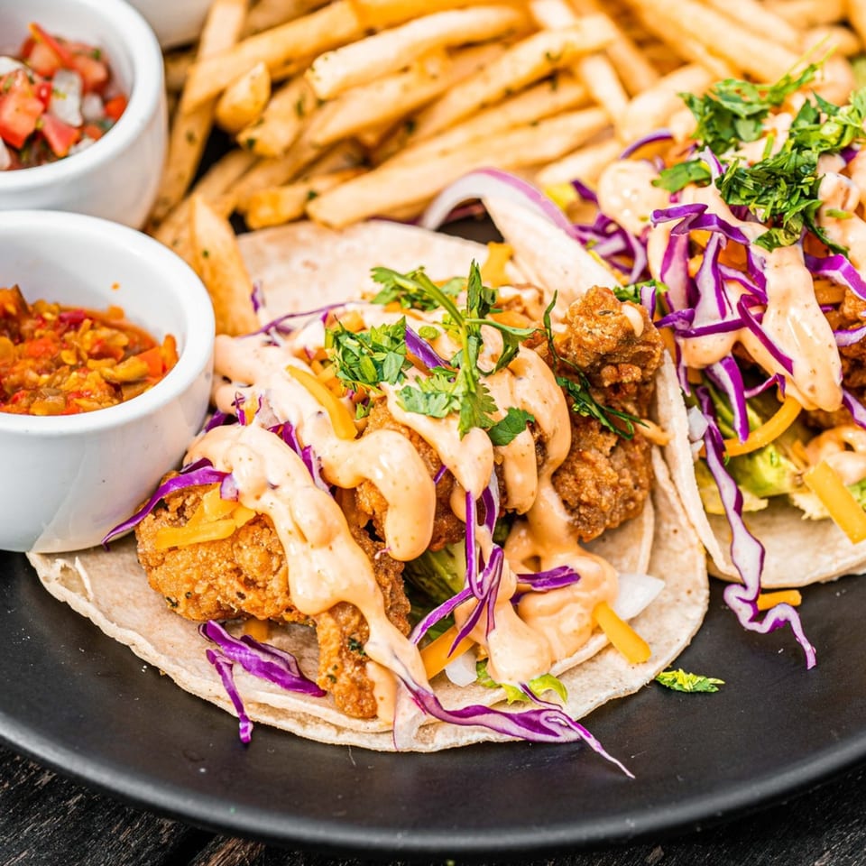 Two Surisan Korean fried chicken tacos with purple cabbage, sauce, and cilantro, served with fries and salsa.