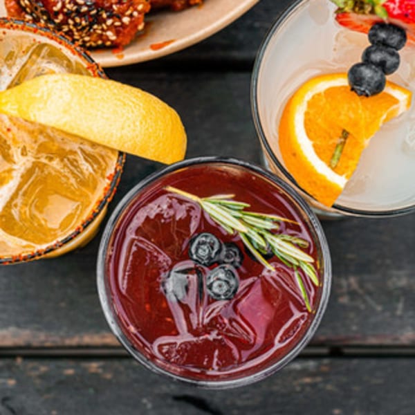 Three vibrant cocktails on a dark wooden table, garnished with citrus, berries, and rosemary.