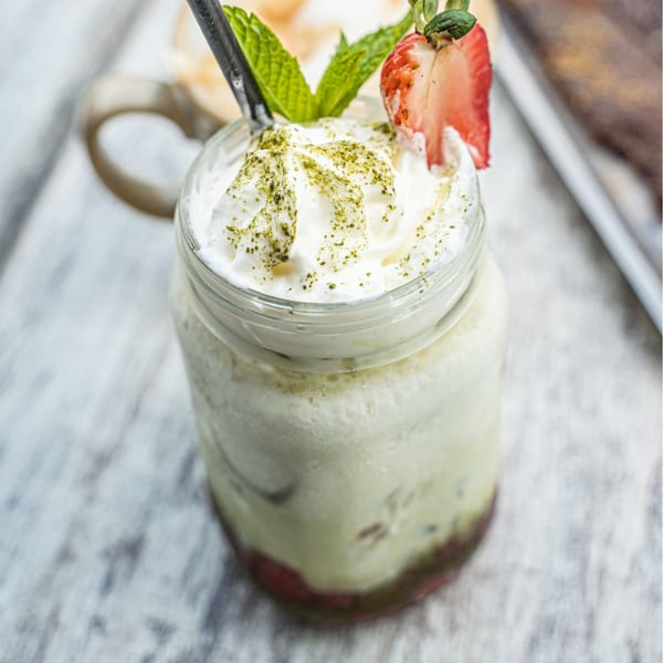 Matcha drink with whipped cream, matcha powder, mint, and a strawberry slice in a mason jar.