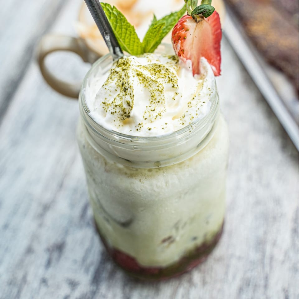 Matcha drink with whipped cream, matcha powder, mint, and a strawberry slice in a mason jar.