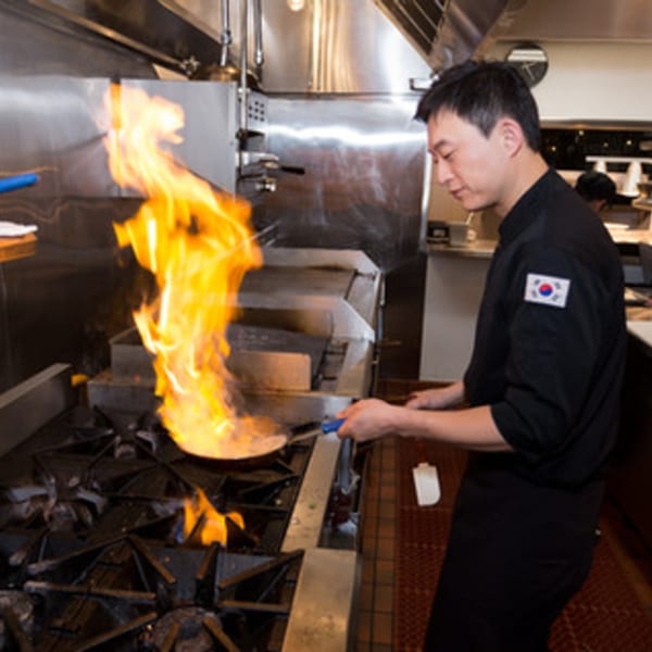 A Surisan chef in a black uniform with a Korean flag patch stir-frying with a large flame.