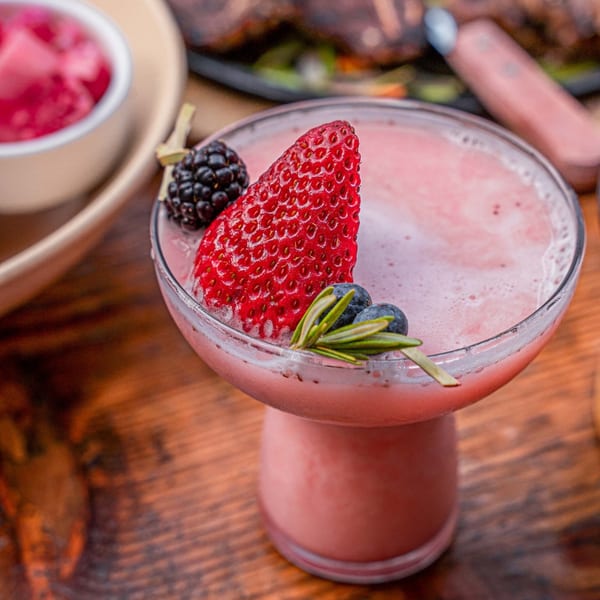A pink cocktail garnished with a large strawberry, blackberries, blueberries, and rosemary sprigs.
