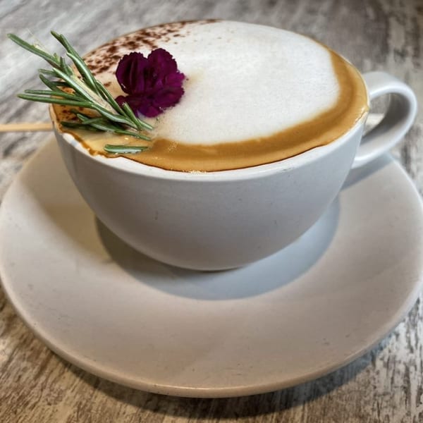 A Surisan latte in a white cup with foam, cocoa powder, a purple flower, and rosemary.