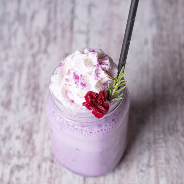 Purple drink with whipped cream, purple sprinkles, a red flower, and rosemary in a glass jar.