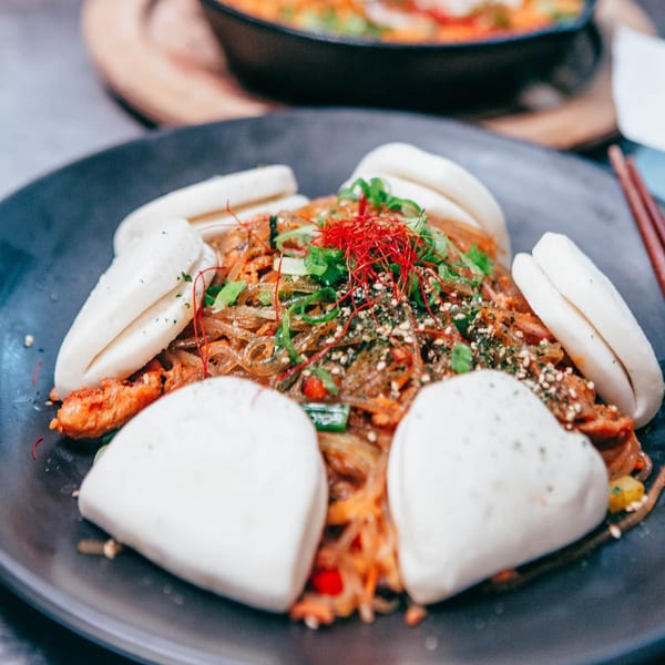 Korean japchae noodles with red chili threads, green onions, sesame seeds, and bao buns.
