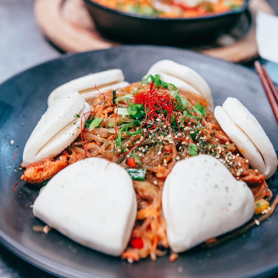 Korean japchae noodles with red chili threads, green onions, sesame seeds, and bao buns.