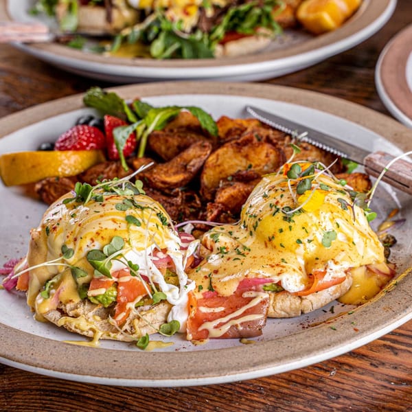Surisan's Eggs Benedict with salmon, avocado, hollandaise, microgreens, potatoes, and fruit on a plate.