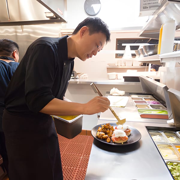 A Surisan chef in a black uniform plates a Korean dish with tongs in a busy kitchen.
